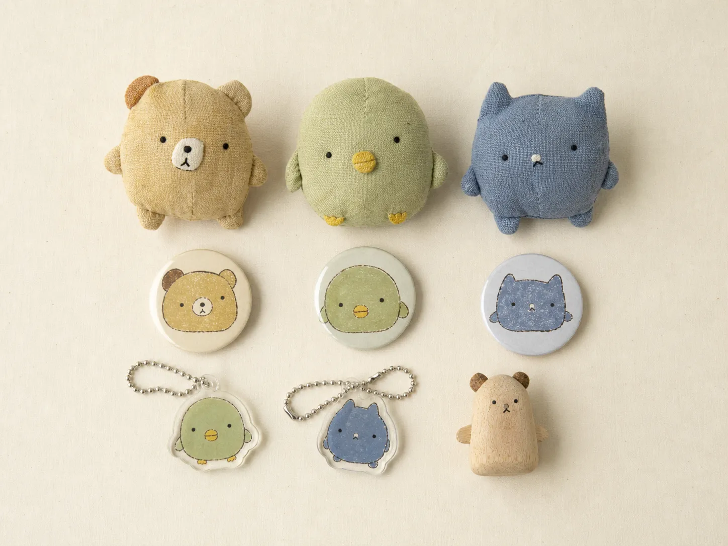 Generated editorial image: souvenir mascot goods on a cream cloth — three palm-sized plush animals in ochre, sage green and slate blue with round bodies, very short limbs and dot eyes, three round tin badges showing the same faces, two small acrylic keychains on ball chains, and a small wooden figure.