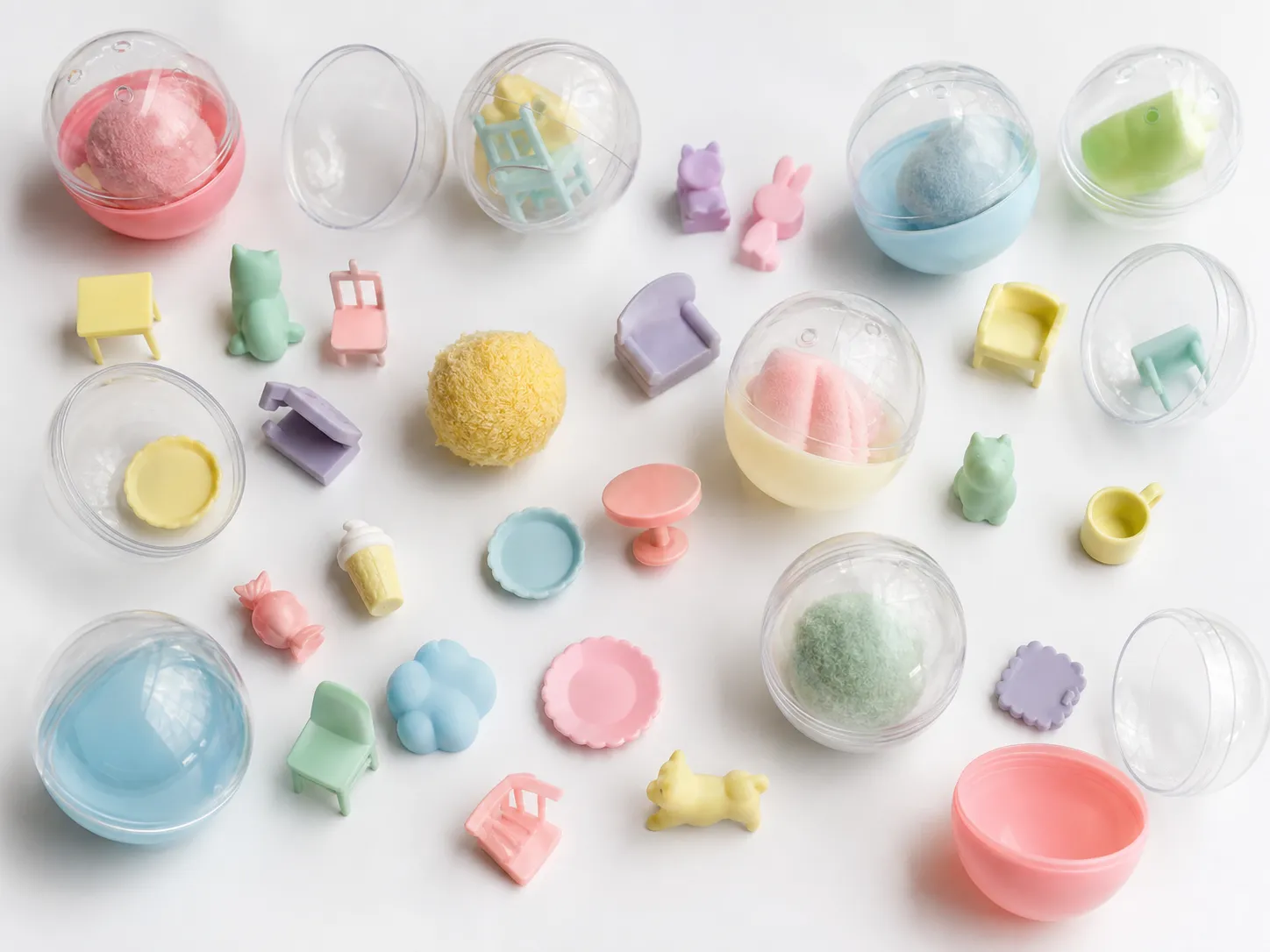 Generated editorial image: clear plastic capsule toy shells, some open, with thumb-sized pastel figures, miniature furniture and small resin objects scattered on a white surface.